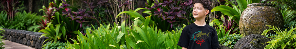 A young boy in a lush tropical garden, wearing a black t-shirt with a colorful graphic print. The background features dense green foliage, purple-leafed plants, a stone wall, and a large decorative pot.
