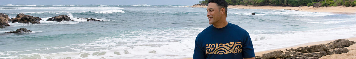 A man wearing a navy blue t-shirt with a gold tribal pattern stands on a rocky beach with the ocean in the background.