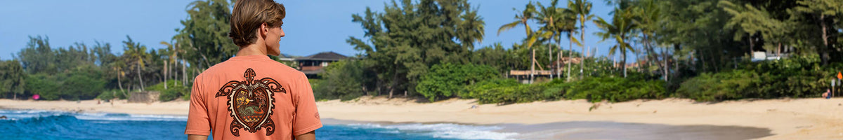 A man standing on a sandy beach facing away from the camera, wearing a coral-orange t-shirt with a large black graphic print on the back. The background features a tropical beach scene with palm trees, blue ocean water, and a clear blue sky.