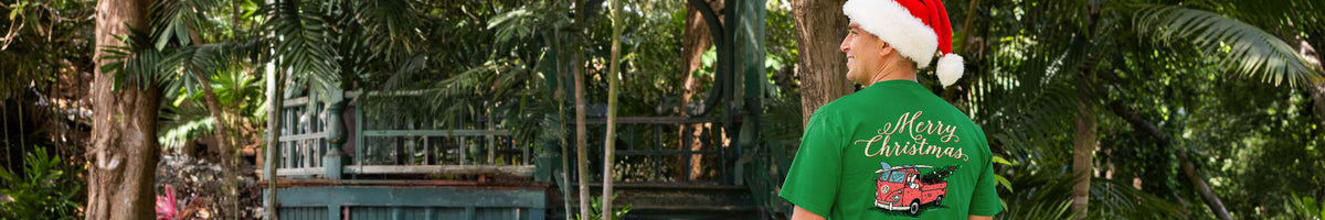 A man wearing a green Christmas-themed t-shirt and a Santa hat, seen from behind in a lush, tropical outdoor setting with trees and a wooden structure.