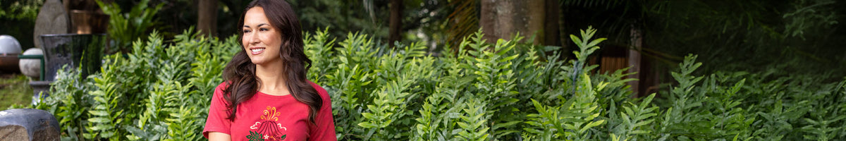 A woman with long dark hair wearing a red top stands outdoors in front of dense green foliage.