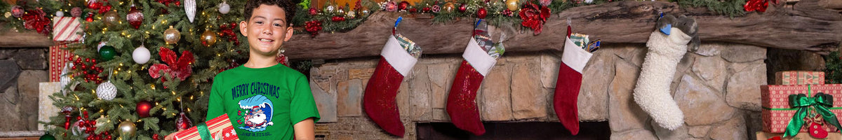 A young boy wearing a green graphic t-shirt stands in front of a decorated Christmas fireplace with stockings, ornaments, and wrapped gifts.