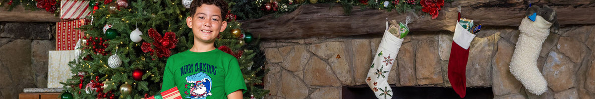 A young boy wearing a green graphic t-shirt stands in front of a decorated Christmas tree and stone fireplace with three stockings hanging from the mantel.
