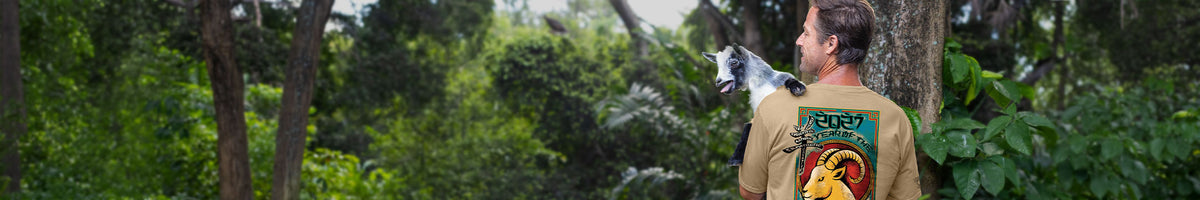 A man wearing a tan t-shirt with a colorful graphic print on the back, standing in a lush green forest and holding a goat.