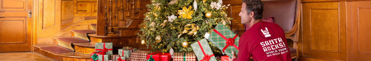 A man in a red long sleeve shirt stands next to a decorated Christmas tree with wrapped gifts on a wooden staircase.