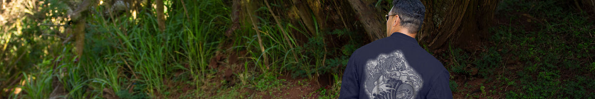 A man viewed from behind wearing a dark navy blue t-shirt with a large white graphic print on the back, standing outdoors in a grassy, wooded environment.