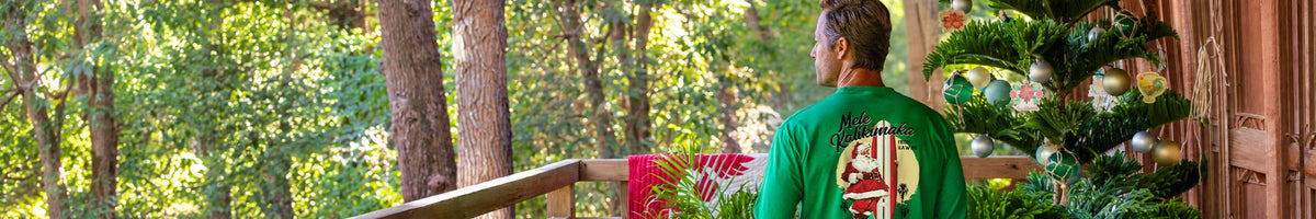 A man viewed from behind wearing a green crewneck t-shirt with a large graphic print on the back, standing on a porch next to a decorated Christmas tree.