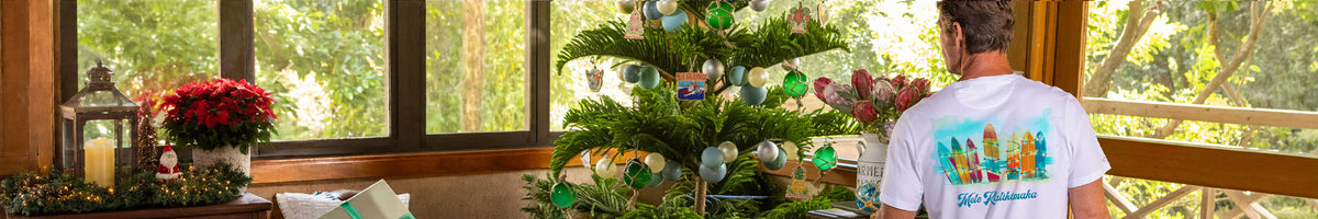 A man wearing a white t-shirt with a colorful graphic and the text 'Mele Kalikimaka' stands in front of a decorated Christmas tree near large windows.
