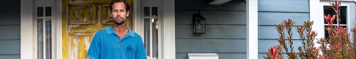 A man wearing a blue polo shirt stands in front of a house porch with blue-gray siding, a yellow door, and red foliage.