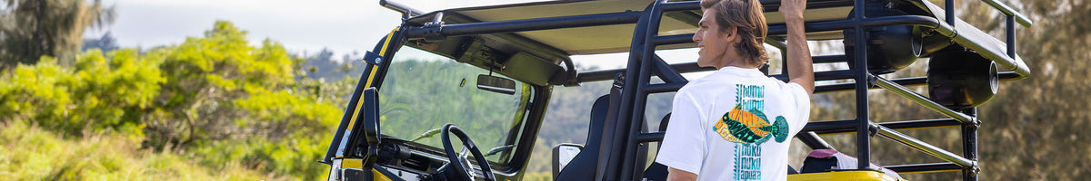 A man wearing a white t-shirt with a colorful fish graphic stands next to a yellow and black off-road vehicle outdoors.