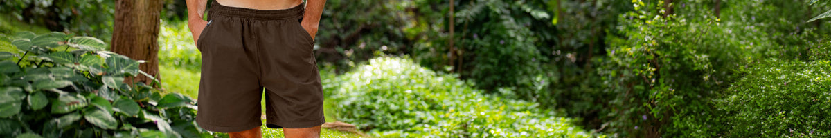 A man wearing brown shorts stands outdoors in a lush green garden or forest setting.