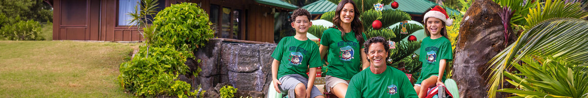 Four people wearing matching green shirts posing together outdoors in front of a tropical house and a decorated Christmas tree.
