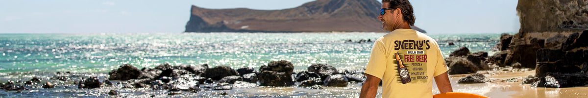 A man wearing a yellow T-shirt with a graphic print stands on a rocky beach looking out toward the ocean and a distant island.