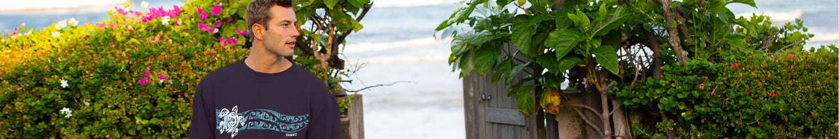 A man wearing a dark navy graphic t-shirt stands outdoors on a beach path with tropical green foliage and the ocean in the background.