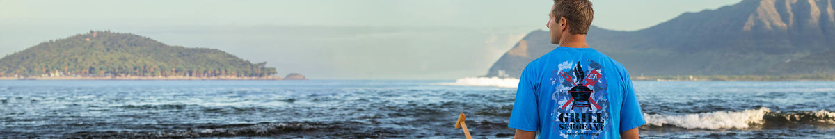 A man standing at the beach facing the ocean, wearing a bright blue t-shirt with a graphic print on the back. The background shows a scenic coastline with mountains and water.
