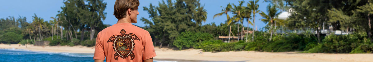 A man standing on a tropical beach with palm trees and blue water, viewed from behind to show a large turtle graphic printed on the back of an orange t-shirt.
