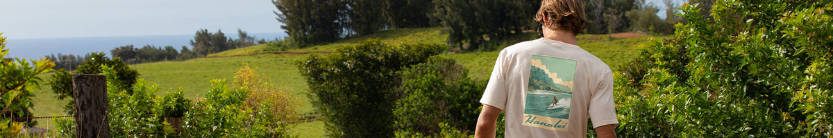 A man viewed from behind wearing a white graphic t-shirt with a green and blue ocean scene print, standing in a lush green outdoor setting with trees and hills.