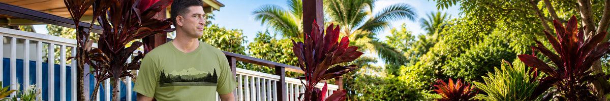 A man wearing a green graphic t-shirt stands outdoors in a tropical garden setting with palm trees and red ornamental plants.