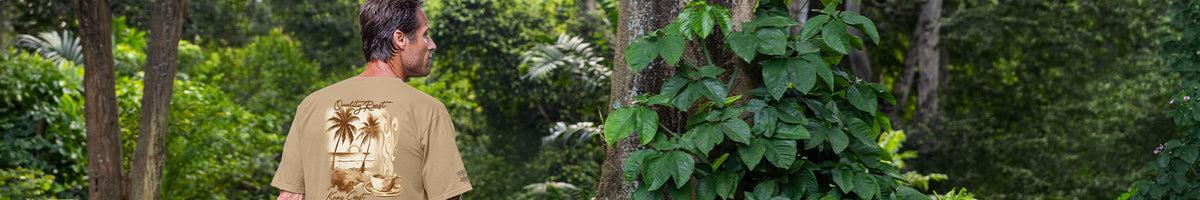 A man viewed from behind wearing a tan t-shirt with a large graphic print of a beach scene on the back, standing in a lush green outdoor setting.