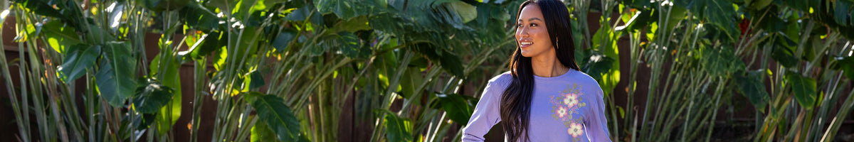 A woman with long dark hair stands outdoors in front of a dense background of green tropical leaves, wearing a light purple long-sleeve top with a white floral graphic on the front.