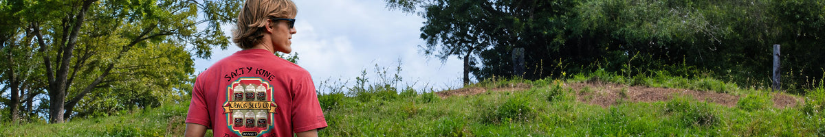 A man wearing a red graphic t-shirt stands on a grassy hill outdoors, looking towards a wooded area under a cloudy sky.