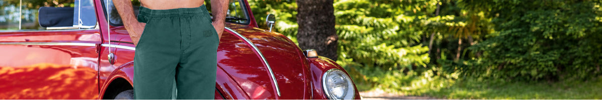 A person wearing dark green trousers stands beside a vintage red car outdoors, with trees and greenery in the background.