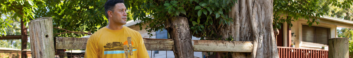 A man wearing a yellow graphic t-shirt stands outdoors in front of a rustic wooden fence and trees.