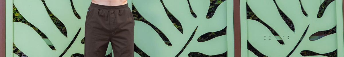 A person wearing dark brown pants stands in front of a mint green decorative wall with black leaf-like cutouts.