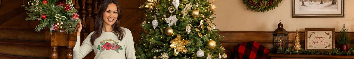 A woman wearing a white Christmas long sleeve with a red and green holiday design stands in a decorated living room next to a lit Christmas tree with gold ornaments.