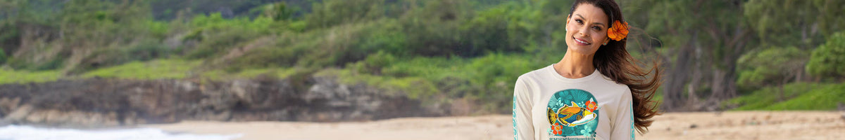 A woman with a brown hair accessory stands on a beach wearing a white long-sleeve t-shirt with a colorful graphic print of a fish, with green forest and cliffs in the background.