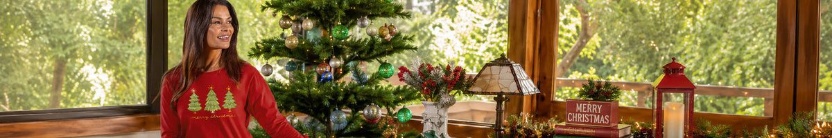 A woman wearing a red Christmas long sleeve with green tree graphics stands indoors near a decorated Christmas tree and warm window light.