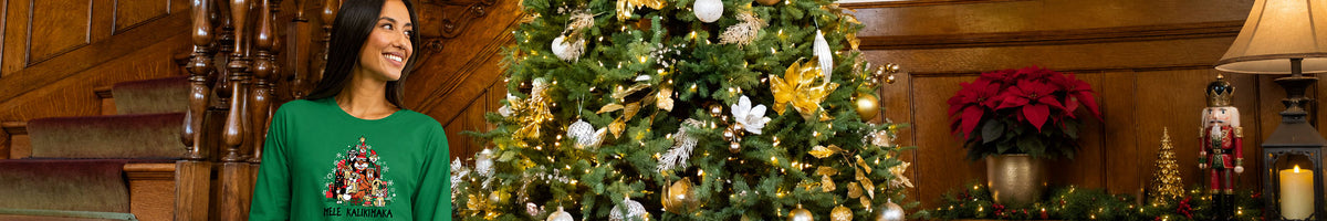 A woman in a green Christmas long sleeve stands next to a decorated Christmas tree in a warm, wood-paneled holiday room with poinsettias and a lamp.
