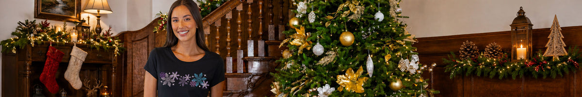 A woman smiling in a dark navy t-shirt with a colorful snowflake pattern, standing in a cozy, decorated holiday home next to a beautifully lit Christmas tree with gold and white ornaments.