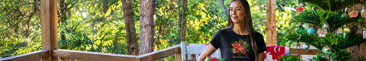 A woman wearing a black graphic t-shirt with a red and green Christmas design, standing in a rustic outdoor porch setting with trees and a decorated Christmas tree in the background.