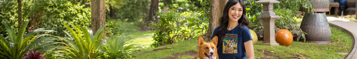 A woman standing outdoors in a garden with a corgi dog, wearing a dark graphic t-shirt. The background features lush green foliage, trees, and a stone lantern.