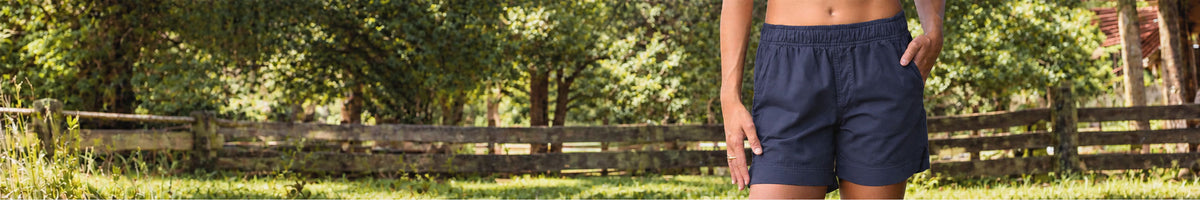 A person outdoors wearing dark purple athletic shorts, standing in a grassy yard with a wooden fence and trees in the background.