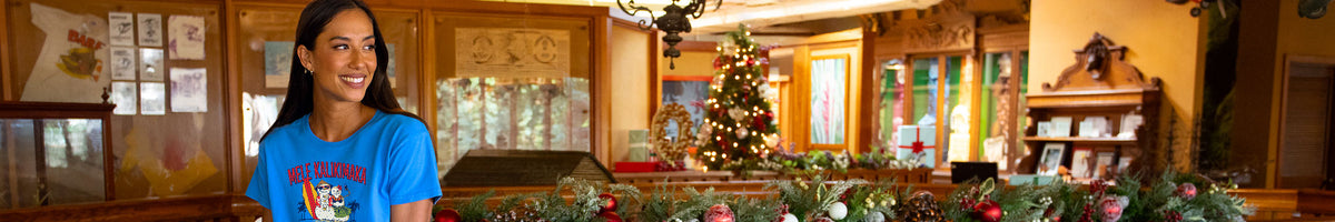 A woman wearing a bright blue graphic t-shirt stands in a rustic indoor setting with wood-paneled walls, a decorated Christmas tree, and holiday garlands in the background.