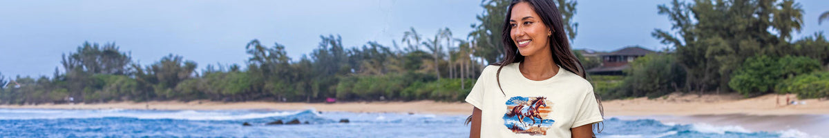 A woman standing on a beach wearing a cream-colored graphic t-shirt with a colorful print on the front. The background shows a tropical beach scene with palm trees, ocean water, and a blue sky.