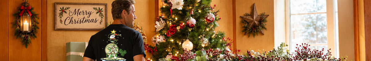 A man wearing a black t-shirt with a Christmas graphic stands in a warmly lit room decorated for Christmas, looking at a decorated tree and a window.