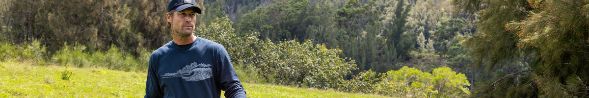 A man wearing a navy blue long-sleeve t-shirt with a tank graphic and a black cap, standing in a grassy field with a forest background.
