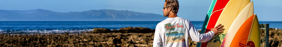 A man on a rocky beach stands with a stack of colorful surfboards, wearing a white long-sleeve t-shirt with a colorful graphic print on the back. The background features a scenic ocean view with distant mountains under a clear blue sky.