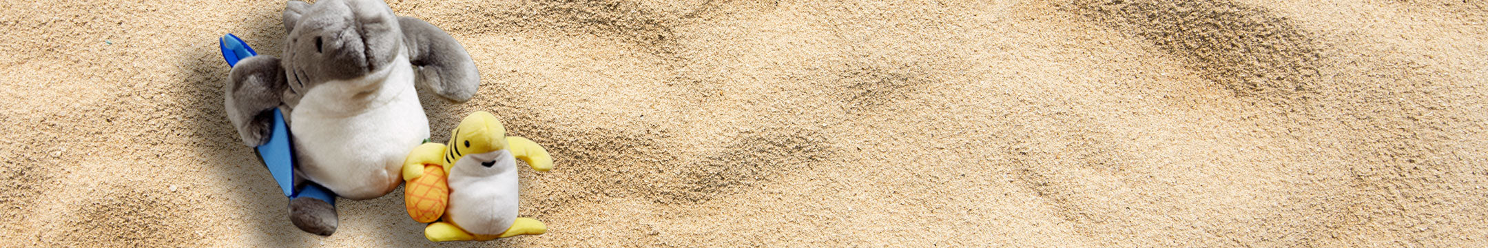 Two plush Sharkas, sitting on sand with a blue surfboards