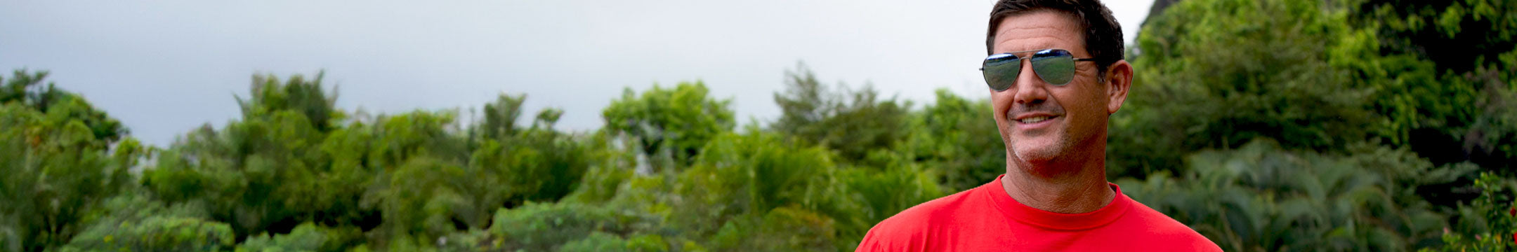 Man wearing sunglasses and a red shirt standing in front of green trees