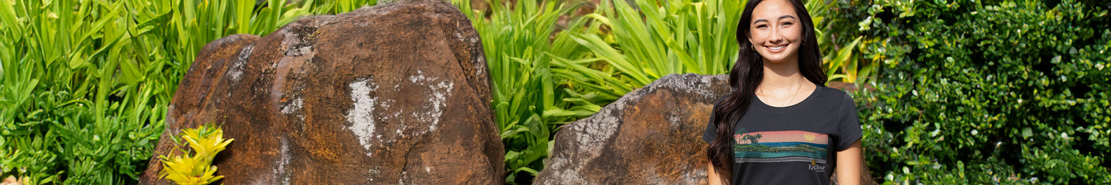 Woman wearing a black shirt with an image of Ko Olina printed on the front standing next to large rocks outside