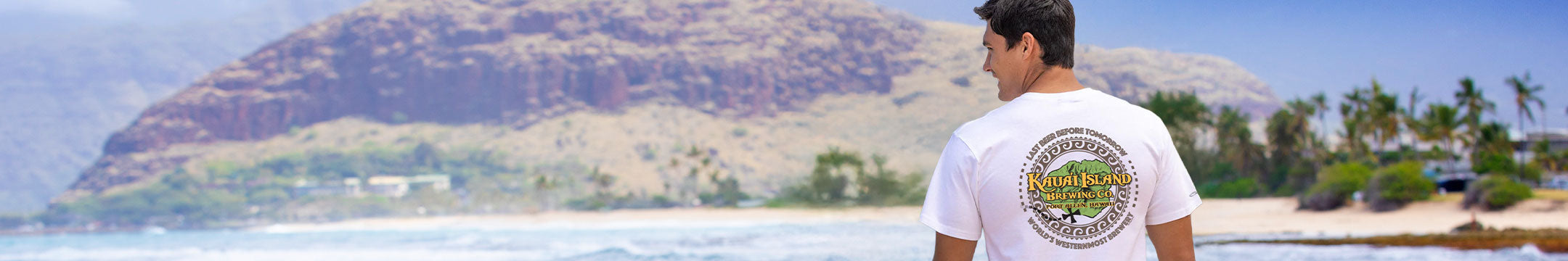 Man wearing a white t-shirt with a Kauai Brewing graphic design, standing on a beach with mountains and ocean in the background.
