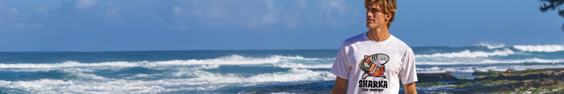 Person wearing a Sharka graphic t-shirt standing on a rocky beach with ocean waves in the background