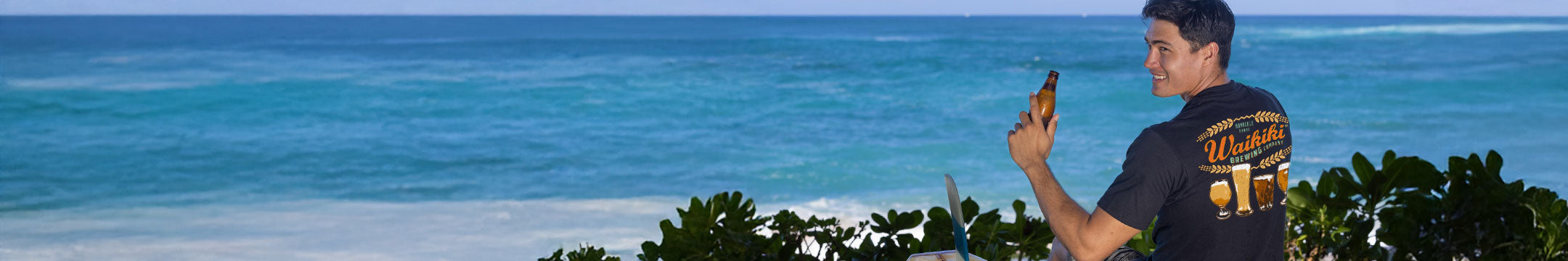 a man drinking a beer on the beach wearing a Waikiki brewing graphic tee
