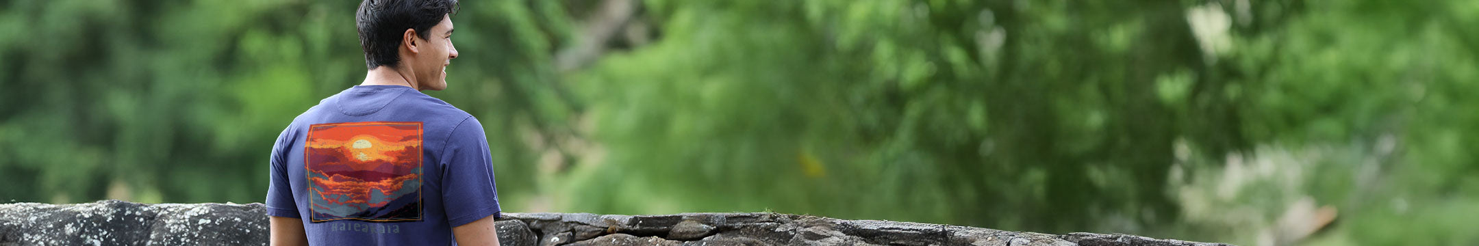 Person wearing a blue t-shirt with a colorful design on the back, standing outdoors with greenery in the background.