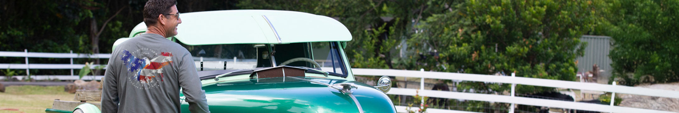Man standing next to a vintage car with a white cover in an outdoor setting wearing a long sleeve shirt with an eagle on it. 