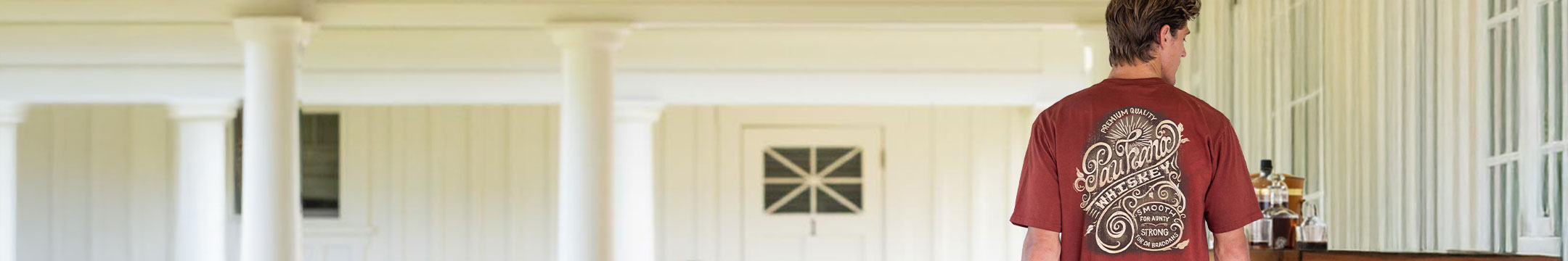 Man standing in a room with white walls and a door, wearing a whiskey dyed shirt with text on the back.
